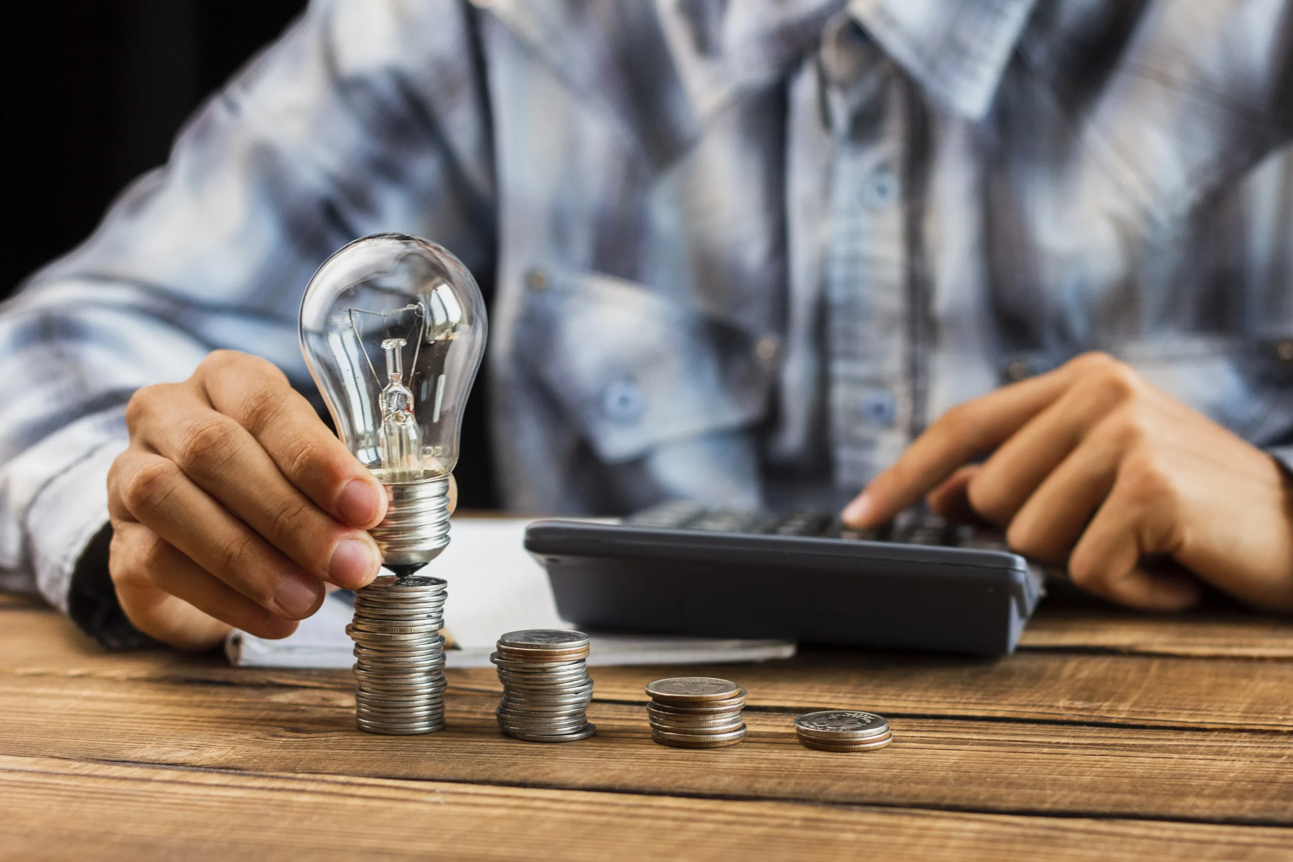 Hand placing a lightbulb on stacked coins beside a calculator, illustrating the decision between storing solar energy or selling it for financial savings.