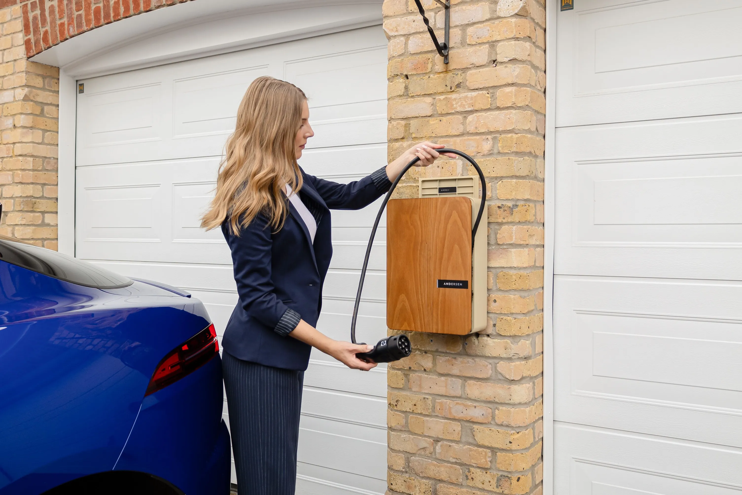 Homeowner plugging an electric vehicle charging cable into a wall-mounted EV charger installed beside a residential garage, illustrating a typical home EV charging setup.