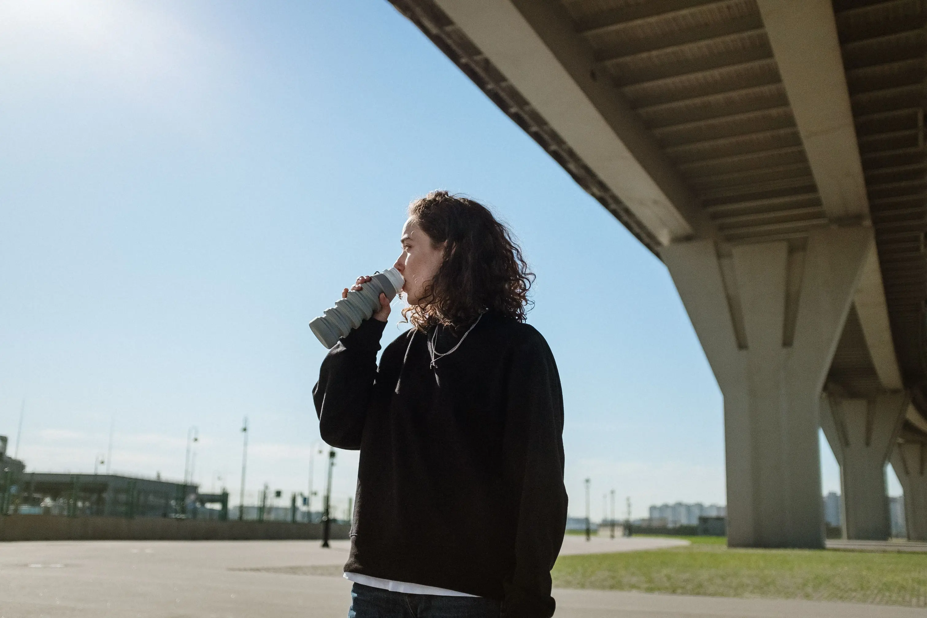 Person drinking from a reusable water bottle beneath an overpass on a sunny day, illustrating the importance of staying hydrated during a UK heatwave while managing household energy use.